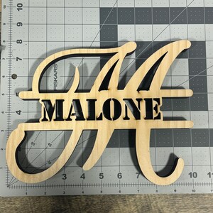 May include: A wooden sign with the letter "M" in a cursive font. The name "MALONE" is cut out of the wood below the letter. The sign is on a gray grid background.