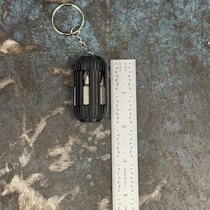 May include: A black multi-tool keychain with a silver key ring and five different screwdriver bits. The tool is about 2 inches long and is sitting on a gray textured surface.
