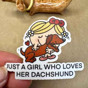 Puede incluir: Una pegatina blanca con una ilustración de dibujos animados de una niña abrazando a un dachshund. El texto "Just a girl who loves her dachshund" está impreso debajo de la ilustración.
