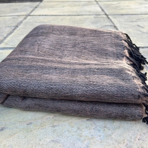 Può includere: Coperta piegata in tessuto marrone e nero con bordo a frange. La coperta ha un aspetto strutturato ed è piegata ordinatamente, mostrando la sua costruzione a strati. La frangia è nera e aggiunge un elemento decorativo.