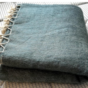 Puede incluir: Una manta de lana verde doblada con un borde de flecos. La manta está hecha de una tela suave y texturizada.