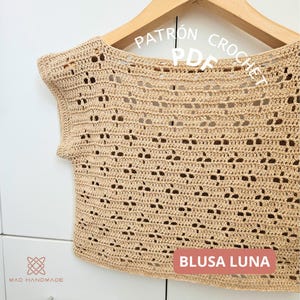 以下が含まれることがあります： ベージュ色の繊細な透かし編みデザインのクロシェ編みトップス。木製のハンガーに掛けられています。画像には「PATRON CROCHET PDF」と「BLUSA LUNA」の文字があります。手作りのトップスは、独特で風通しの良いパターンが特徴です。