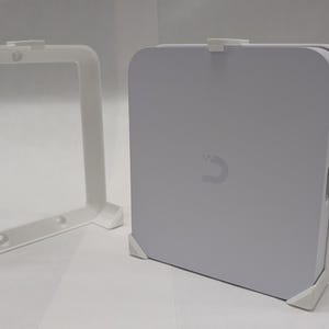 May include: White plastic wall mount for a small device. The mount has two brackets that attach to the wall and a center piece that holds the device. The device is not included.