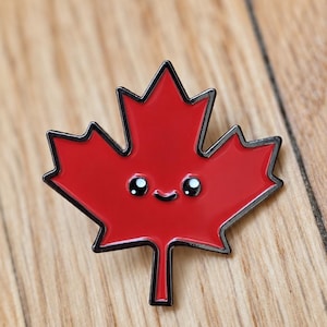 Puede incluir: Un broche de esmalte con forma de hoja de arce roja y contorno de metal negro. La hoja presenta un rostro sencillo y tierno con dos pequeños ojos negros con reflejos blancos y una fina línea negra curvada que forma una sonrisa.