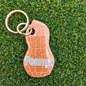 May include: A gold key chain with a peanut-shaped charm. The charm is brown and has a textured surface with the word "Peanut" printed in blue.