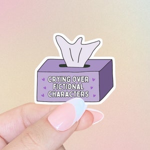 Puede incluir: Una pegatina de caja de pañuelos de color morado con el texto "Crying over fictional characters" y pequeños corazones alrededor del texto.