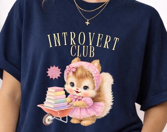 May include: Navy blue t-shirt with the text "Introvert Club" in a vintage font. The shirt features a cartoon squirrel wearing a pink dress and headscarf, pushing a wheelbarrow of books. A pink starburst graphic is also present.