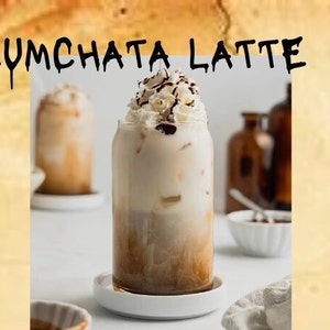 Puede incluir: Un vaso de Rumchata Latte con crema batida y un chorrito de chocolate. La bebida está en un vaso transparente con un posavasos blanco. El fondo es una imagen borrosa de una superficie marrón.