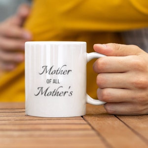Può includere: Tazza in ceramica bianca con la scritta "Mother of All Mother's" in un carattere corsivo. La tazza ha linee verticali ed è tenuta da una persona. Lo sfondo è sfocato, con un tavolo di legno in primo piano.