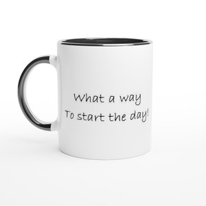 May include: White ceramic mug with a black rim and handle. The mug has the text "What a way to start the day!" in black script. The mug is set against a white background.