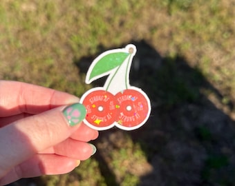 May include: A cherry sticker with a green leaf and stem. The two red cherries have the words "STRONG AF" printed on them. The sticker is held in a hand against a blurred background of grass.