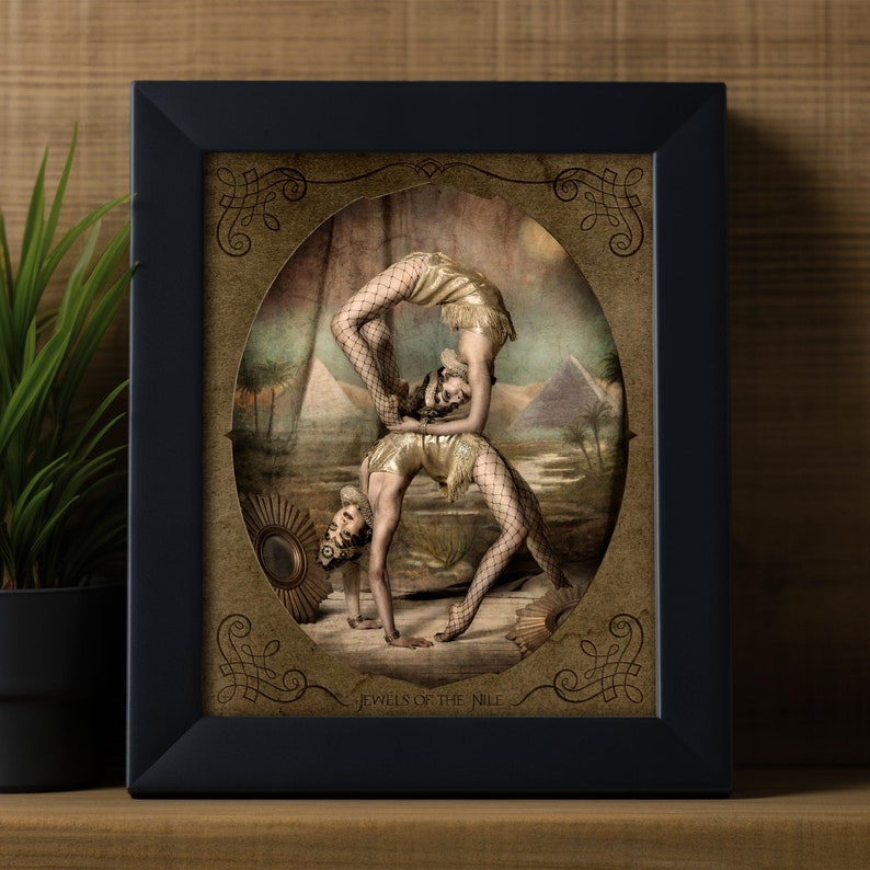 May include: Two women in gold and black costumes perform an acrobatic pose in front of a backdrop depicting an Egyptian landscape. The image is framed in a black frame with a gold oval border and the text "Jewels of the Nile".