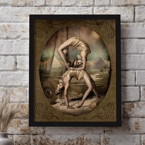 May include: A vintage-style framed print featuring two women performing an acrobatic pose in front of a desert landscape with pyramids. The print has a gold background with ornate borders and the text "Jewels of the Nile".