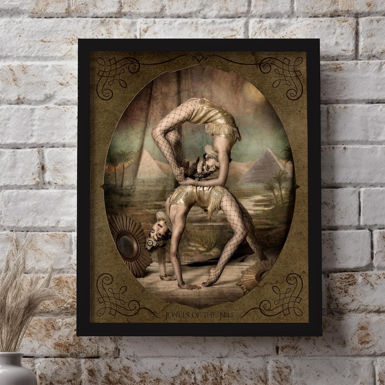 May include: A vintage-style framed print featuring two women performing an acrobatic pose in front of a desert landscape with pyramids. The print has a gold background with ornate borders and the text "Jewels of the Nile".