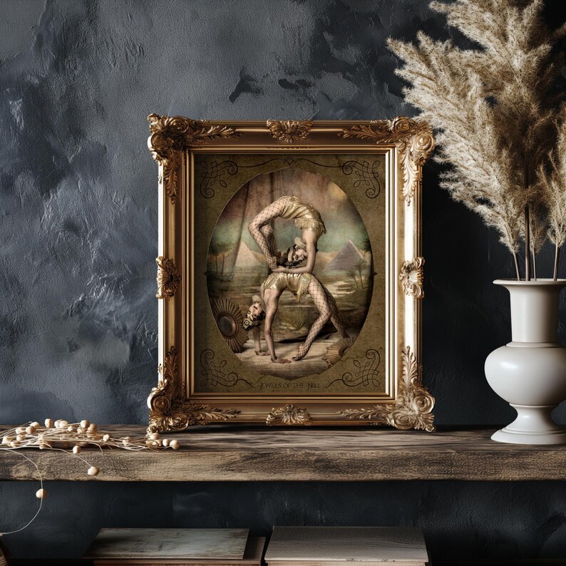 May include: A framed print of two women performing an acrobatic pose in front of a desert landscape. The print is in a gold frame with ornate details. The text "Queens of the Nile" is at the bottom of the print.