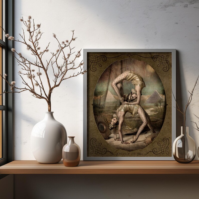 May include: A framed print of two women performing an acrobatic pose. The women are wearing vintage-style clothing and are set against a backdrop of a desert landscape. The print is titled "Jewels of the Nile".