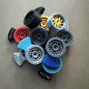 Może przedstawiać: Zbiór 10 różnych kolorowych i wzorzystych kół do samochodu zdalnie sterowanego. Koła są wykonane z tworzywa sztucznego i mają różne wzory szprych. Koła są oznaczone tekstem "TY.T RC".