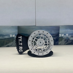 Puede incluir: Una rueda plateada impresa en 3D con una base negra y un logotipo TY.T RC negro. La rueda está diseñada para un coche de control remoto.
