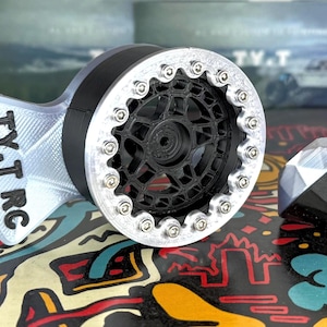 以下が含まれることがあります： 銀と黒の3Dプリントされたおもちゃの車のホイールで、黒いタイヤと銀のリムが付いています。ホイールは詳細なデザインで、「TY-T RC」とラベル付けされています。