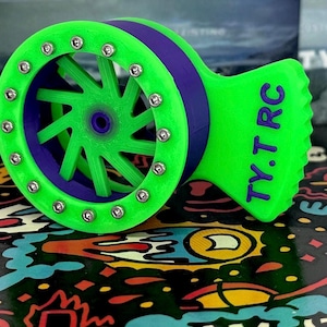 Może przedstawiać: Koło 3D drukowane w kolorze zielonym i fioletowym z wzorem gwiazdy i srebrnymi śrubami wokół obręczy. Koło ma napis "TY.T.RC" nadrukowany na boku.