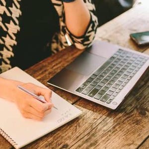 May include: A person is writing in a notebook with a pen while sitting at a wooden table with a laptop and a cell phone.