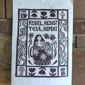 May include: A white print on paper with a brown ink design. The design includes the words "REBEL, RESIST, rest, REPEAT" and a woman in a mug. Floral and geometric elements surround the central image. The print is mounted on a brick wall.