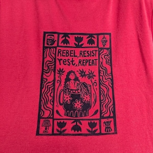 Pode incluir: Camiseta vermelha com estampa gráfica preta. A estampa apresenta as palavras "REBEL, RESIST, REST, REPEAT" em torno de uma imagem central de uma mulher e uma caneca floral. O design é emoldurado por elementos decorativos.