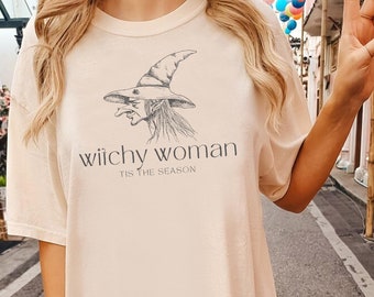 May include: Cream-colored t-shirt featuring a witch illustration and the text "Wiichy Woman, Tis The Season" in a black font. The witch is depicted wearing a pointed hat. The shirt has short sleeves and a relaxed fit.