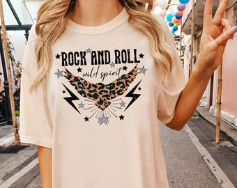 May include: A cream-colored t-shirt with a graphic of a black and brown leopard print eagle with the text "Rock and Roll wild spirit" and black lightning bolts and stars.