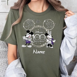 May include: A green t-shirt with a silver glitter design featuring Mickey and Minnie Mouse celebrating Disney's 100th anniversary. The design includes the text "Disney 1923 2023 100 Years of Wonder" and a customizable name field.
