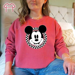 Peut inclure: Un sweat-shirt rose avec un cercle &agrave; carreaux noirs et blancs autour d'une image de dessin anim&eacute; de Mickey Mouse.