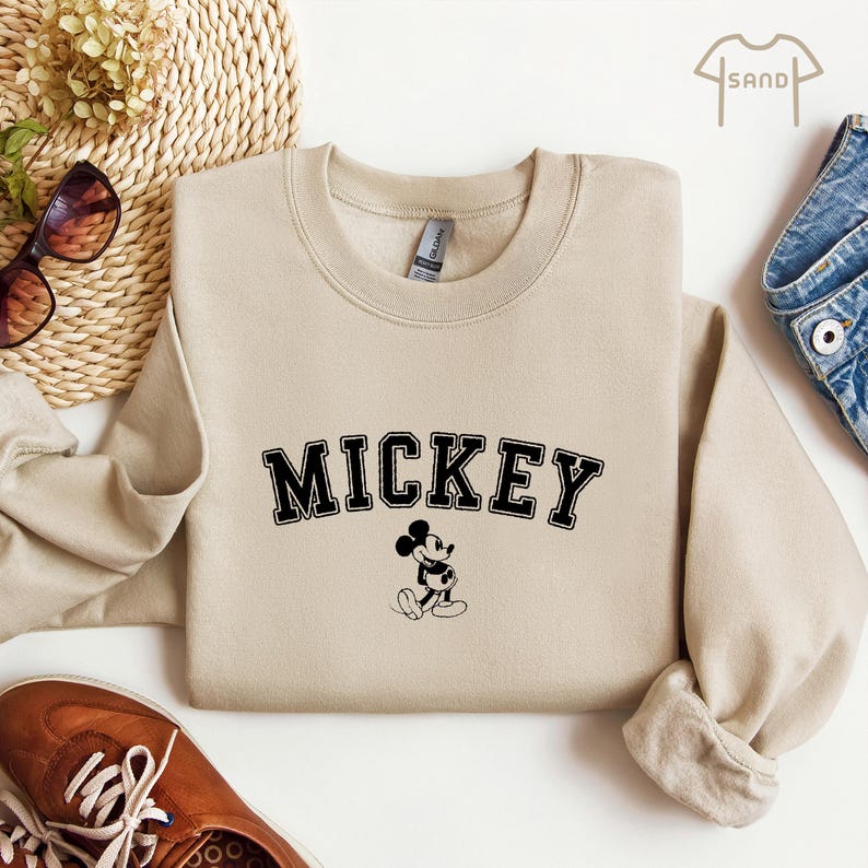 Puede incluir: Una sudadera beige con un gr&aacute;fico de Mickey Mouse negro y la palabra "MICKEY" en letras negras.