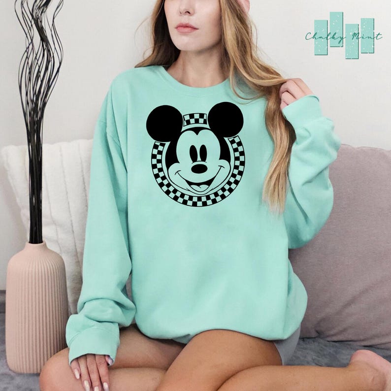 Peut inclure: Un sweat-shirt vert clair avec un cercle &agrave; carreaux noir et blanc autour d'une image noir et blanc du visage de Mickey Mouse.