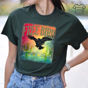 May include: Dark green t-shirt with a graphic featuring a black eagle with outstretched wings, the words "FREE BIRD" in a colorful rectangle, and the word "AMERICA" below. Two green cacti are also featured.