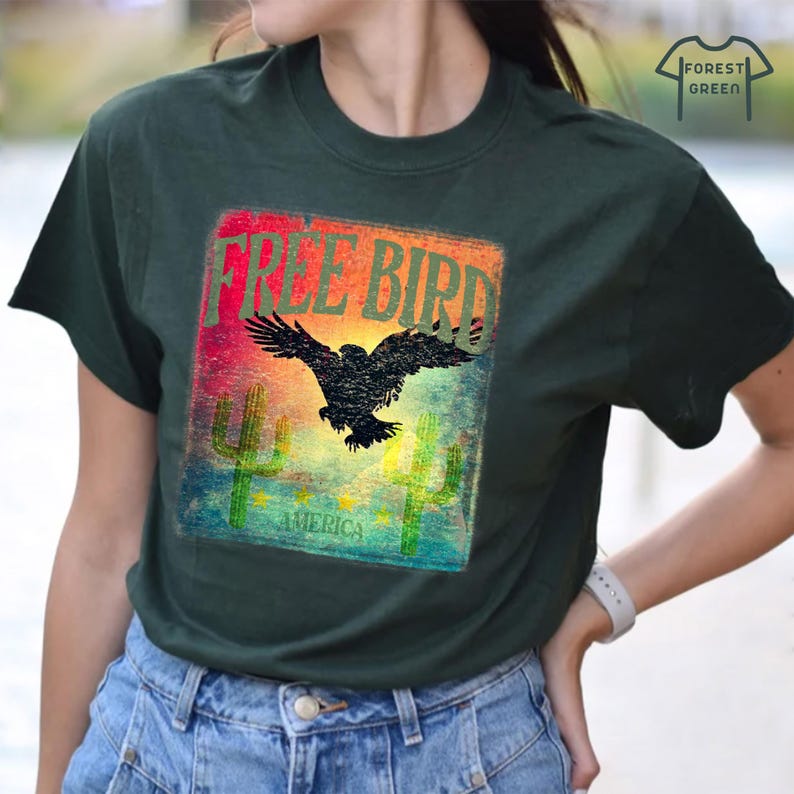 May include: Dark green t-shirt with a graphic featuring a black eagle with outstretched wings, the words "FREE BIRD" in a colorful rectangle, and the word "AMERICA" below. Two green cacti are also featured.