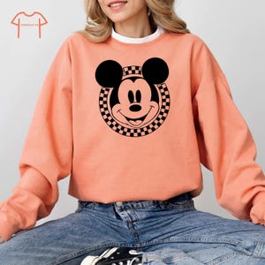 Peut inclure: Un sweat-shirt couleur p&ecirc;che avec un motif circulaire noir et blanc &agrave; carreaux repr&eacute;sentant le visage de Mickey Mouse.
