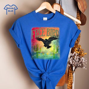 May include: Royal blue t-shirt with a graphic featuring a black eagle with outstretched wings, the words "FREE BIRD" and "AMERICA", and cactus illustrations. The design has a vintage, distressed look.