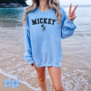 Puede incluir: Una sudadera de color azul claro con la palabra "MICKEY" impresa en negro y una imagen en blanco y negro de Mickey Mouse. La sudadera la lleva puesta una persona de pie en una playa.