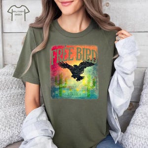 May include: Military green t-shirt with a graphic featuring a black bird silhouette, the words "FREE BIRD" in red and orange, and the word "AMERICA" below. The design includes cactus illustrations and a colorful background.
