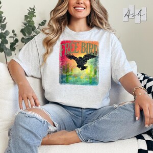 May include: A light gray t-shirt featuring a graphic with the words "FREE BIRD" above a black eagle silhouette. The graphic has a colorful sunset background with cactus illustrations and the word "AMERICA" below.