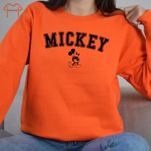 Puede incluir: Sudadera naranja con texto negro que dice "MICKEY" y una silueta negra de Mickey Mouse.