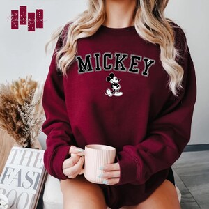 Puede incluir: Una sudadera color burdeos con la palabra "MICKEY" impresa en negro y un gr&aacute;fico de Mickey Mouse en blanco y negro debajo.