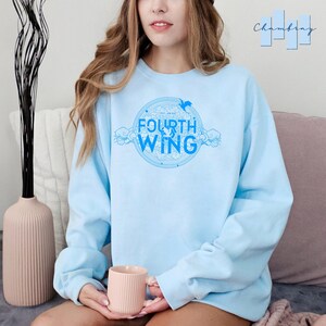 May include: A light blue sweatshirt with a graphic design of a circle with the words "The Law of" and "Fourth Wing" inside. The graphic also includes a bird and clouds.