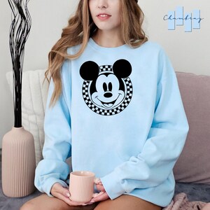 Peut inclure: Un sweat-shirt bleu clair avec un motif circulaire noir et blanc &agrave; carreaux repr&eacute;sentant le visage de Mickey Mouse.