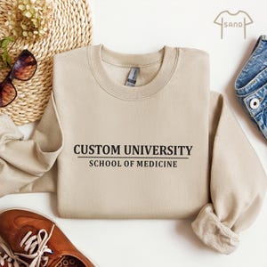 May include: A beige sweatshirt with the text "CUSTOM UNIVERSITY SCHOOL OF MEDICINE" in black. The sweatshirt is folded, with a pair of sunglasses, a woven basket, and a pair of brown shoes in the background.