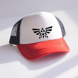 May include: A red and white trucker hat with a black mesh back. The hat features a black graphic of a stylized emblem on the front. The brim is red, and the crown is white. A stylish accessory for fans of video games.
