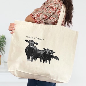May include: Cream-colored tote bag featuring a black and white illustration of three cows. The text "thank a farmer" is printed above the cows. The bag has a simple design with a rectangular shape and long straps.