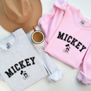 Puede incluir: Dos sudaderas, una gris y otra rosa, con un gr&aacute;fico de Mickey Mouse negro y la palabra "MICKEY" en una fuente universitaria negra.