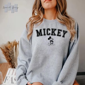 Puede incluir: Una sudadera gris con la palabra "MICKEY" impresa en letras negras y una imagen en blanco y negro de Mickey Mouse.
