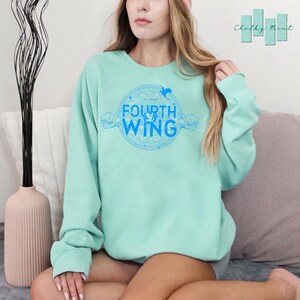 May include: A light blue sweatshirt with a blue graphic design of a circle with the words "The Fourth Wing" and a bird inside the circle.